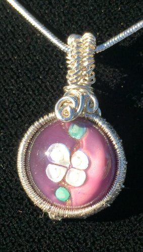 A purple 14mm blown glass cabochon depicting an apple blossom is woven into a fine silver pendant, then set on a 20