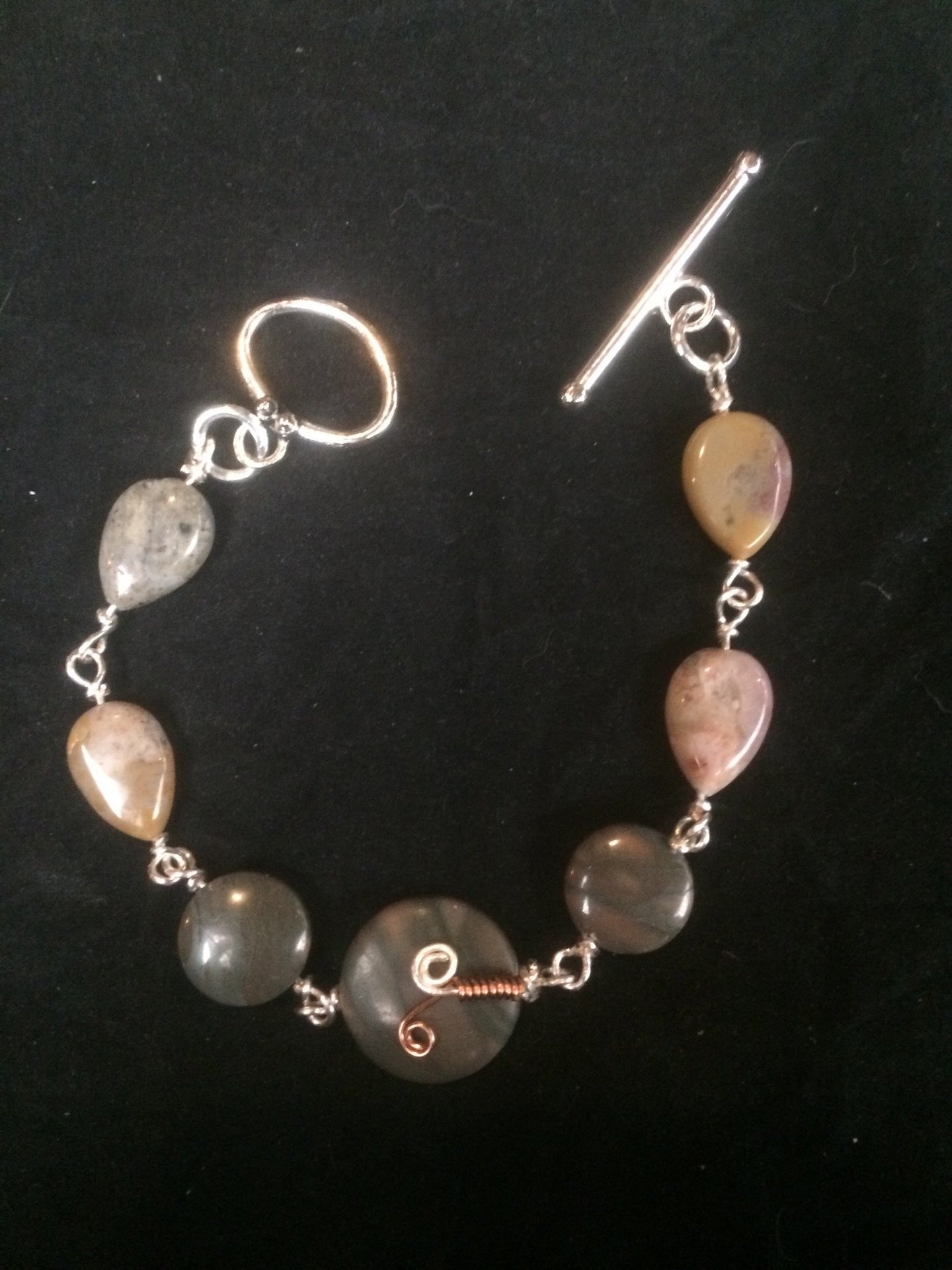 Four 15x10mm teardrop shaped agate beads lead away from two 13mm agate disk beads and an 18mm focal agate bead adorned with a copper sprout wire wrap pattern. A matching silver plated 20x13mm oval toggle clasp completes this 8