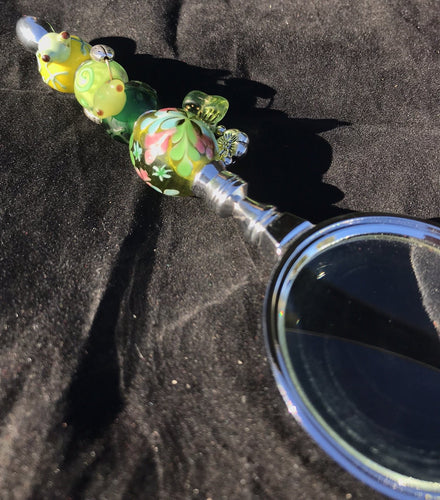 A blown glass bead depicting a tiny frog face peeking out from underwater caps off the sunlit garden imagery on the handle of this magnifying glass.