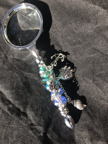 An anchor charm fixes the location conjured up by the sea life charms and dazzling blue-green cut and blown glass beads on the handle of this magnifying glass.