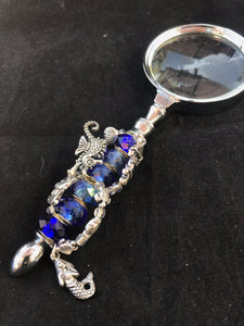 A mermaid charm is enthroned atop a swirl of sea life and nautical charms that dive and bob around a "water column" of dazzling blue glass beads on the handle of this magnifying glass.