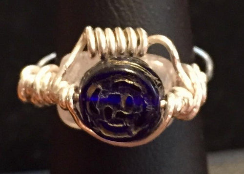 A natural 10x8mm cherry quartz bead is wrapped behind an embossed blue glass bead with fine silver wire coils in this fanciful ring. Rings made by this wire wrapping method vary individually. This one is approxmately a size 4.75, but may fit a slightly larger finger depending on how snug a fit you prefer.